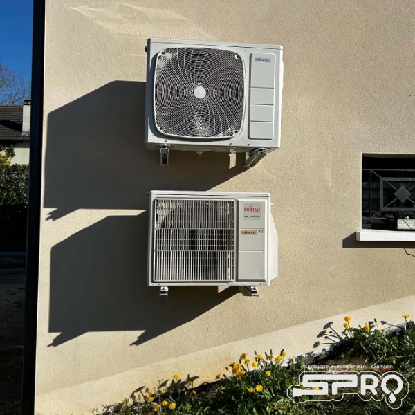 Sur un mur en crépis se trouve deux unités de clim d'extérieur, à côté d'une petite fenêtre.