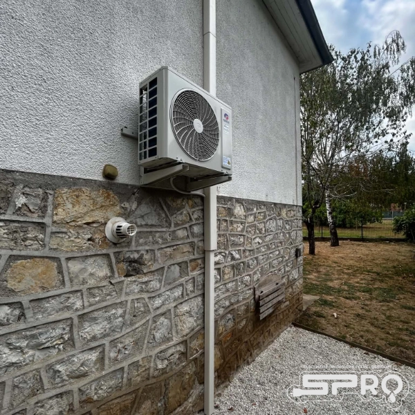 Unité de climatisation extérieur sur mur mi-pierre, mi-crépis. Une ventilation se trouve au dessous et derrière la climatisation se trouve les gaines pour les câbles électriques. Sur le côté gauche de la photo se trouve le jardin du client.