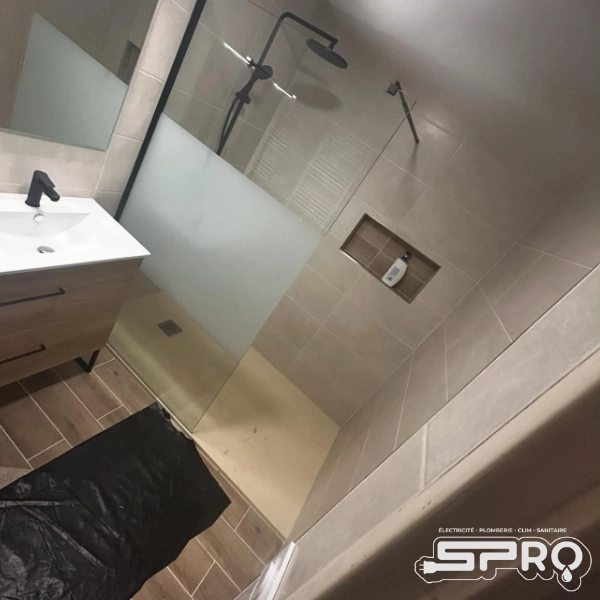 Salle de bain rénover au couleur beige-rose. On y voit un évier sur meuble en bois et un miroir au dessus. Derrière se trouve une vitre, séparant alors le reste de la douche. Le mur est creuser dans la douche pour pouvoir déposer les produits. Une bâche noire se trouve sur le sol, devant l'évier.