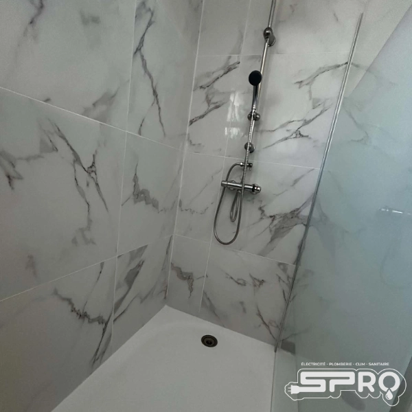Douche au carrelage imitation marbre, une vitre sur le côté droit de l'image et le bac de douche est blanc.