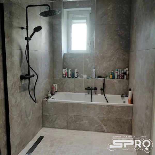 Un espace de salle de douche où se trouve à l'arrière une baignoire sous une fenêtre lumineuse. Le carrelage est rosé et la tuyauterie est noire.